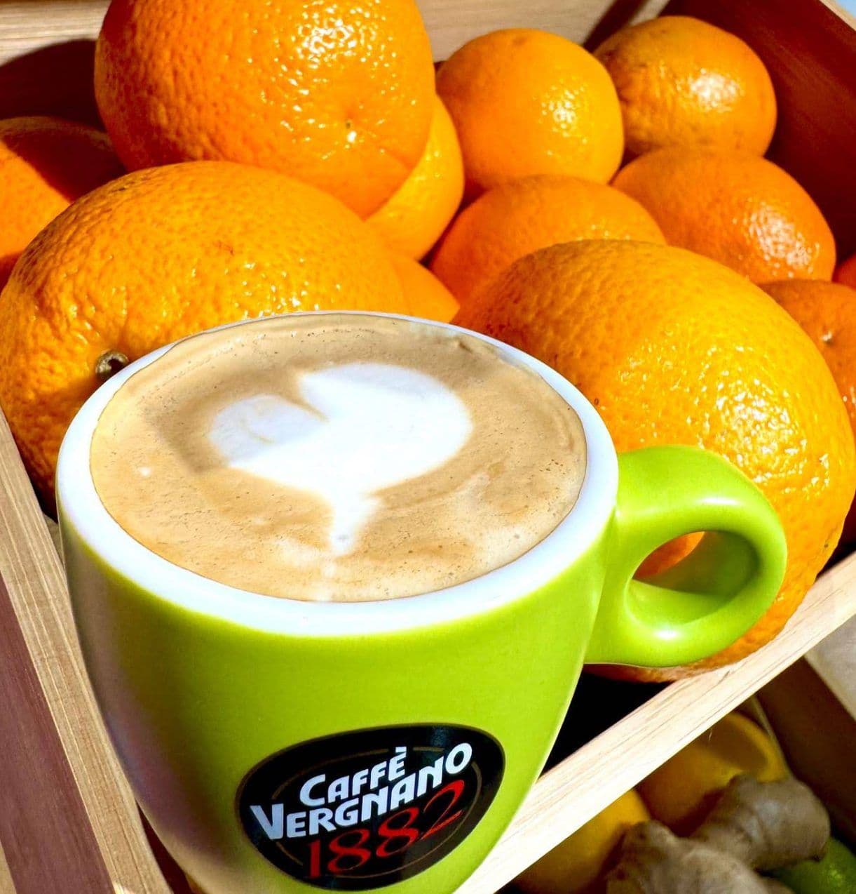 Cappuccino într-o ceașcă verde, în fața unei lădițe cu portocale